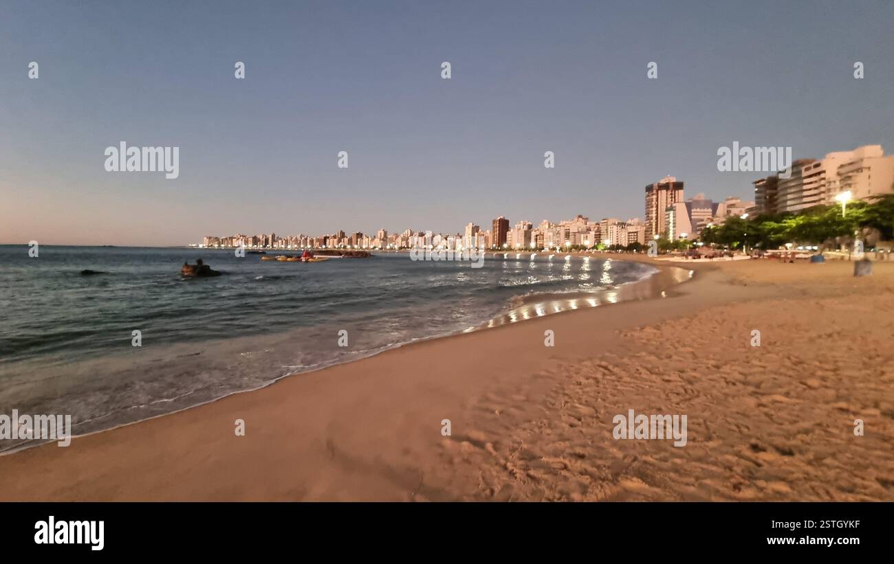 As the sun begins to set, the golden lights of the city reflect beautifully on the calm ocean waters, creating a mesmerizing glow along the shoreline. - Smartphone Captured Stock Image