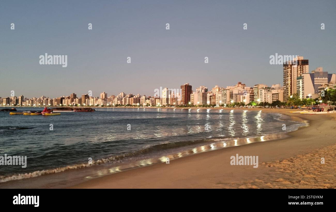 As the sun begins to set, the golden lights of the city reflect beautifully on the calm ocean waters, creating a mesmerizing glow along the shoreline. - Smartphone Captured Stock Image