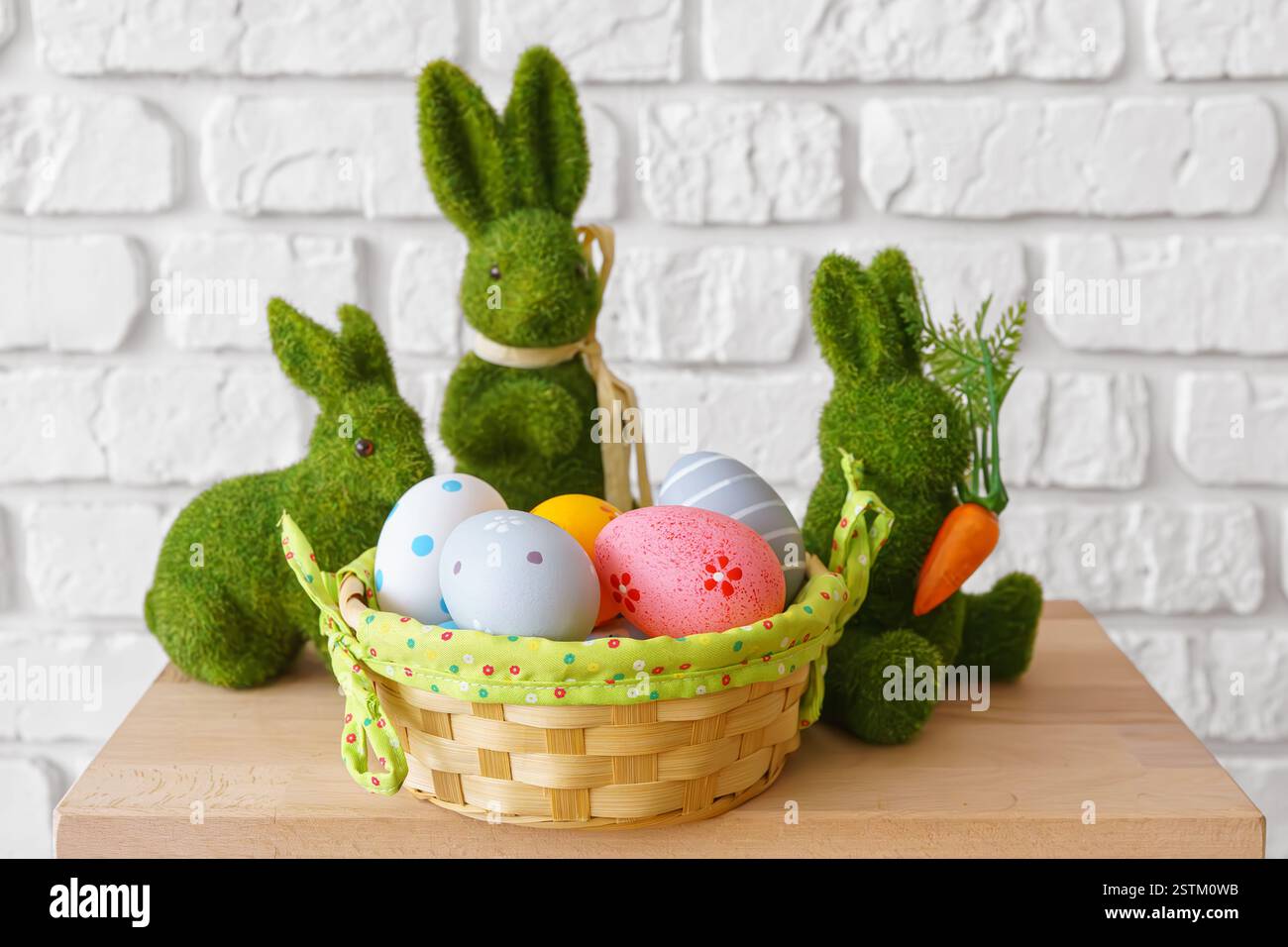 Trhee decorative green moss bunny figures with colorful Easter eggs in wicker basket on wooden table on white brick wall background. Easter holiday fe Stock Photo