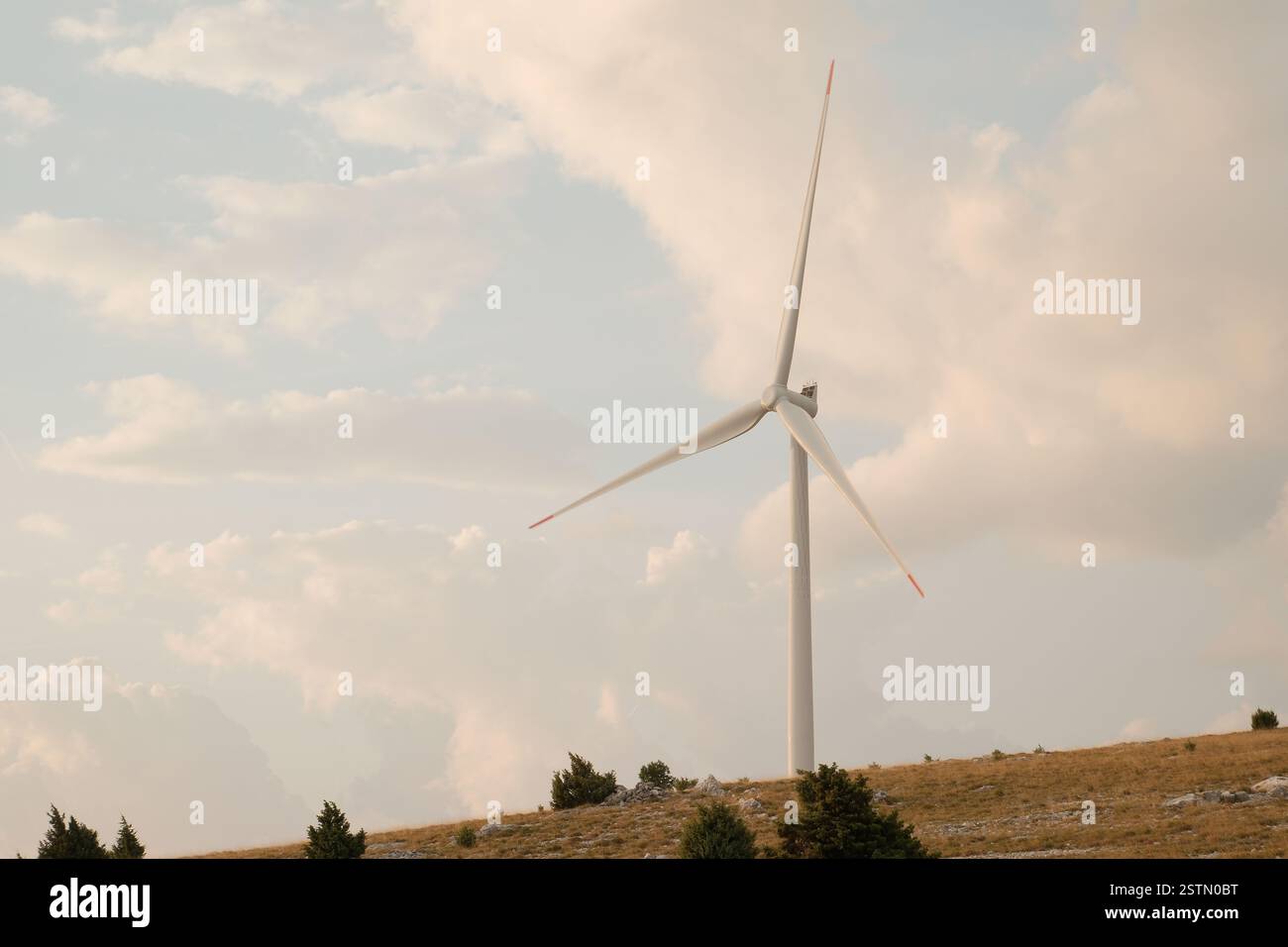 A single wind turbine stands tall against a partly cloudy sky atop a hill Stock Photo