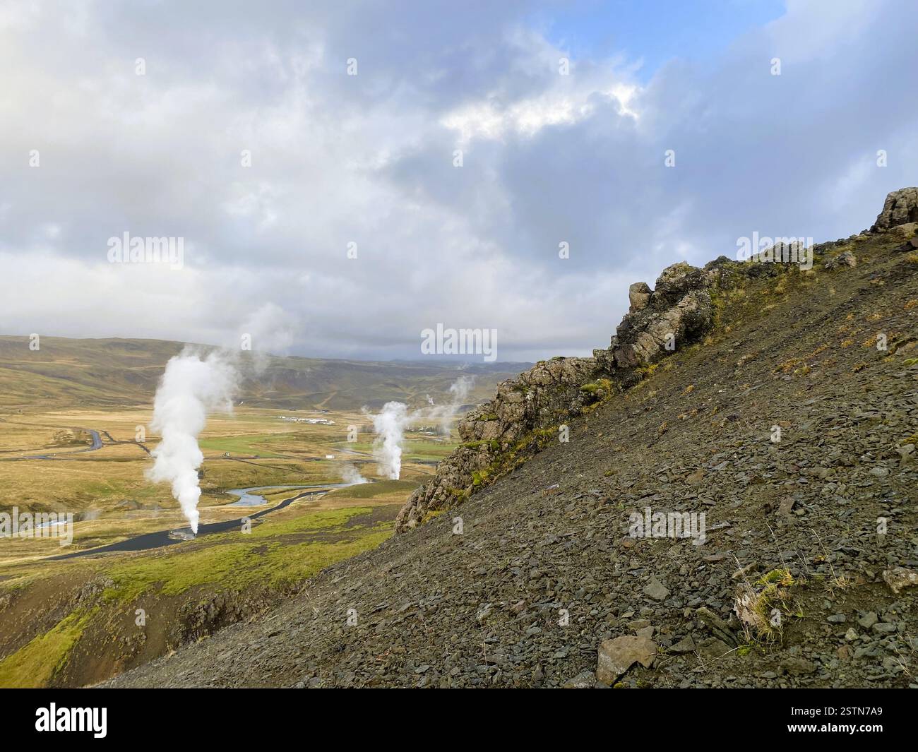 The photo features a rugged landscape with dramatic steam vents, a lush valley, and a winding river reflecting the sky, geothermal activity Stock Photo