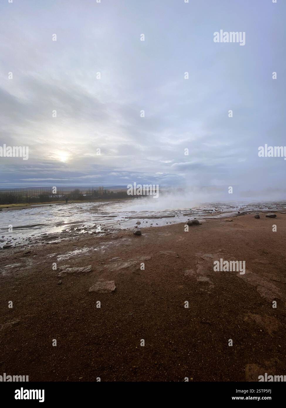 A rugged geothermal landscape with reddish-brown earth, steam rising from the surface, and a cloudy sky, creating an otherworldly atmosphere Stock Photo