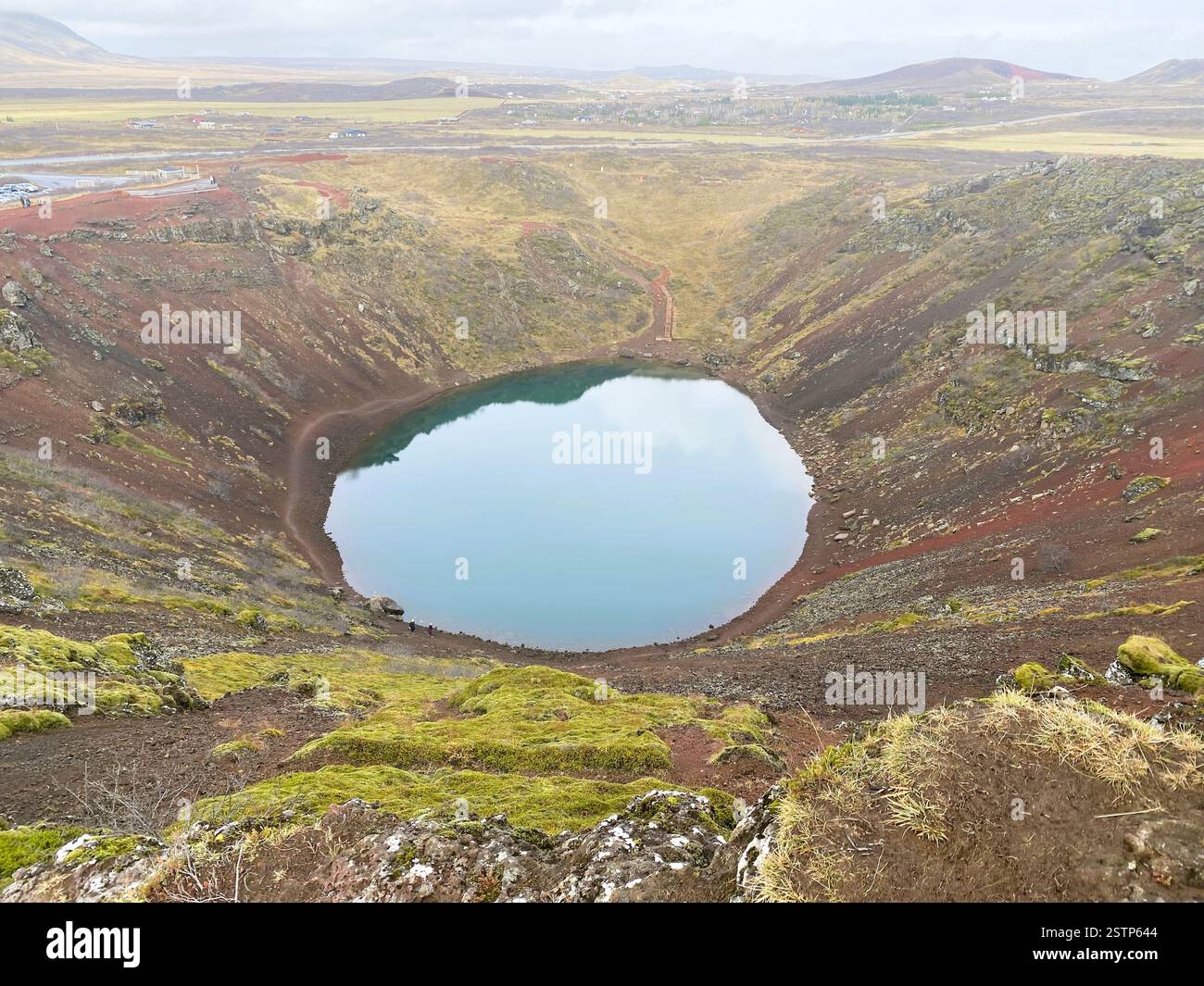 A stunning volcanic crater lake with circular shape and blue water, surrounded by steep, rugged walls of dark volcanic rock and patches of green moss Stock Photo
