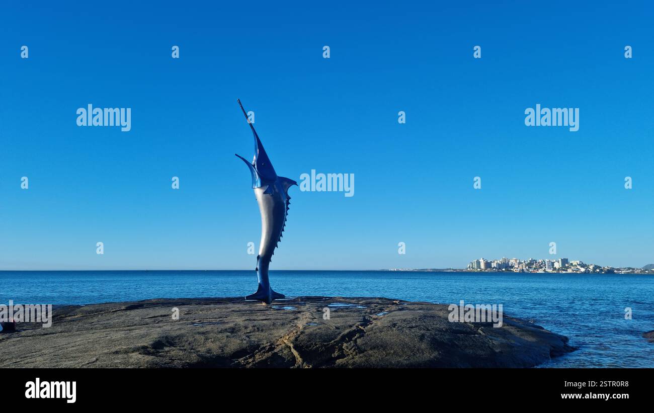 This striking image features a towering marlin sculpture standing proudly on a rocky shoreline, appearing as if it is leaping out of the sea. - Smartphone Captured Stock Image