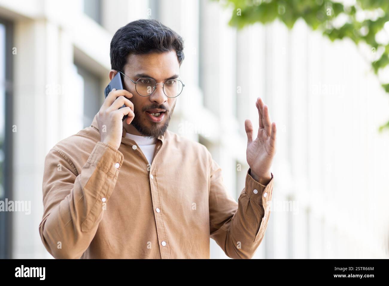 A young man with a beard wearing eyeglasses is having an argument on the phone outdoors with an angry expression and waving his hand in frustration Stock Photo