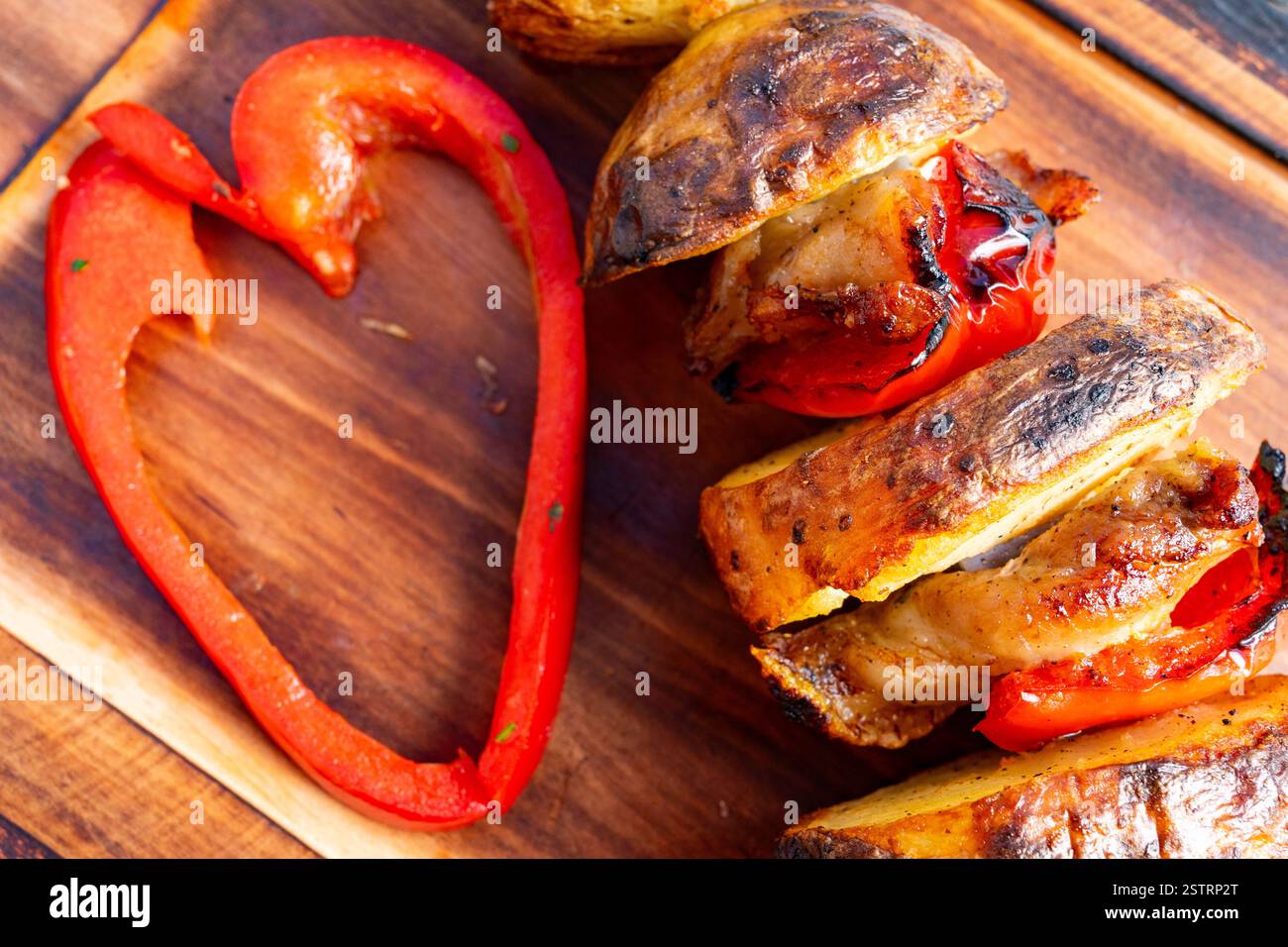 Juicy chicken skewers paired with roasted vegetables are presented on a wooden board. A creative touch is added with a heart-shaped slice of red peppe Stock Photo