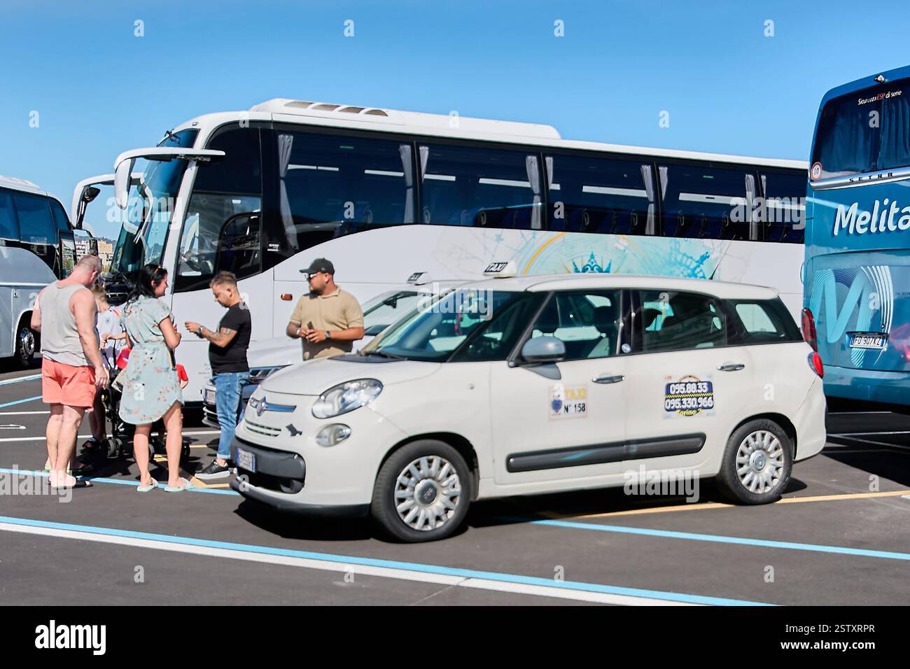 Catania. Italy - February 20, 2025: A white taxi and a tourist bus parked in Catania, Sicily, with people conversing nearby. The image captures a typi Stock Photo