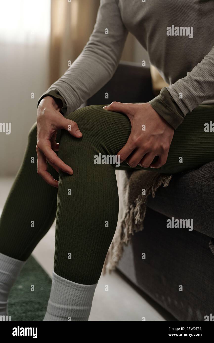 Person sitting on grey couch while stretching arm and shoulder. Soft light coming through window creating a peaceful atmosphere in cozy living room Stock Photo