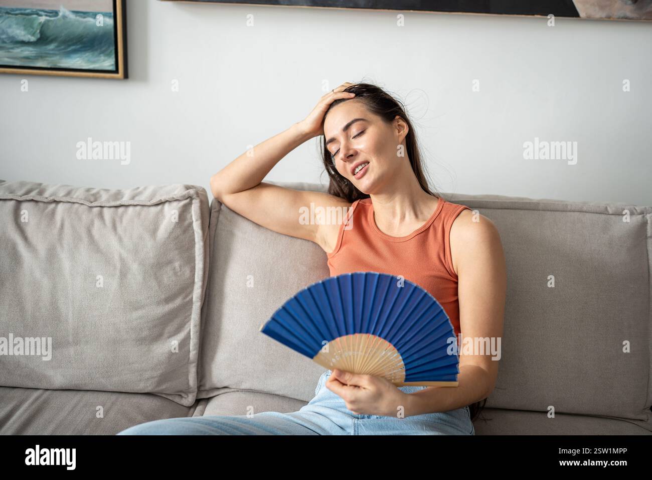 Glad woman successfully coping with abnormal heat, blowing with paper fan, sitting on couch. Stock Photo