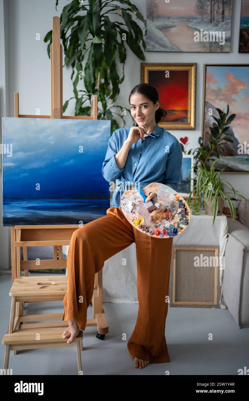 Portrait artist woman stands near easel with canvas in art studio hold colour palette look at camera Stock Photo