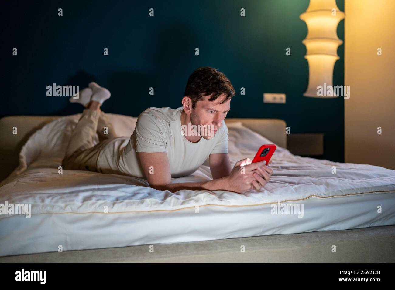 Man reading information in smartphone lying in bed at home in modern bedroom in evening. Stock Photo