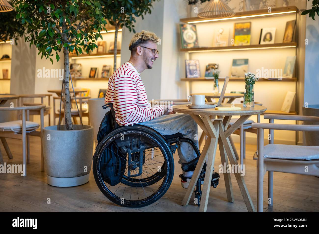 Happy pleased man on wheelchair in cafe using laptop for communication, video call, online chatting. Stock Photo