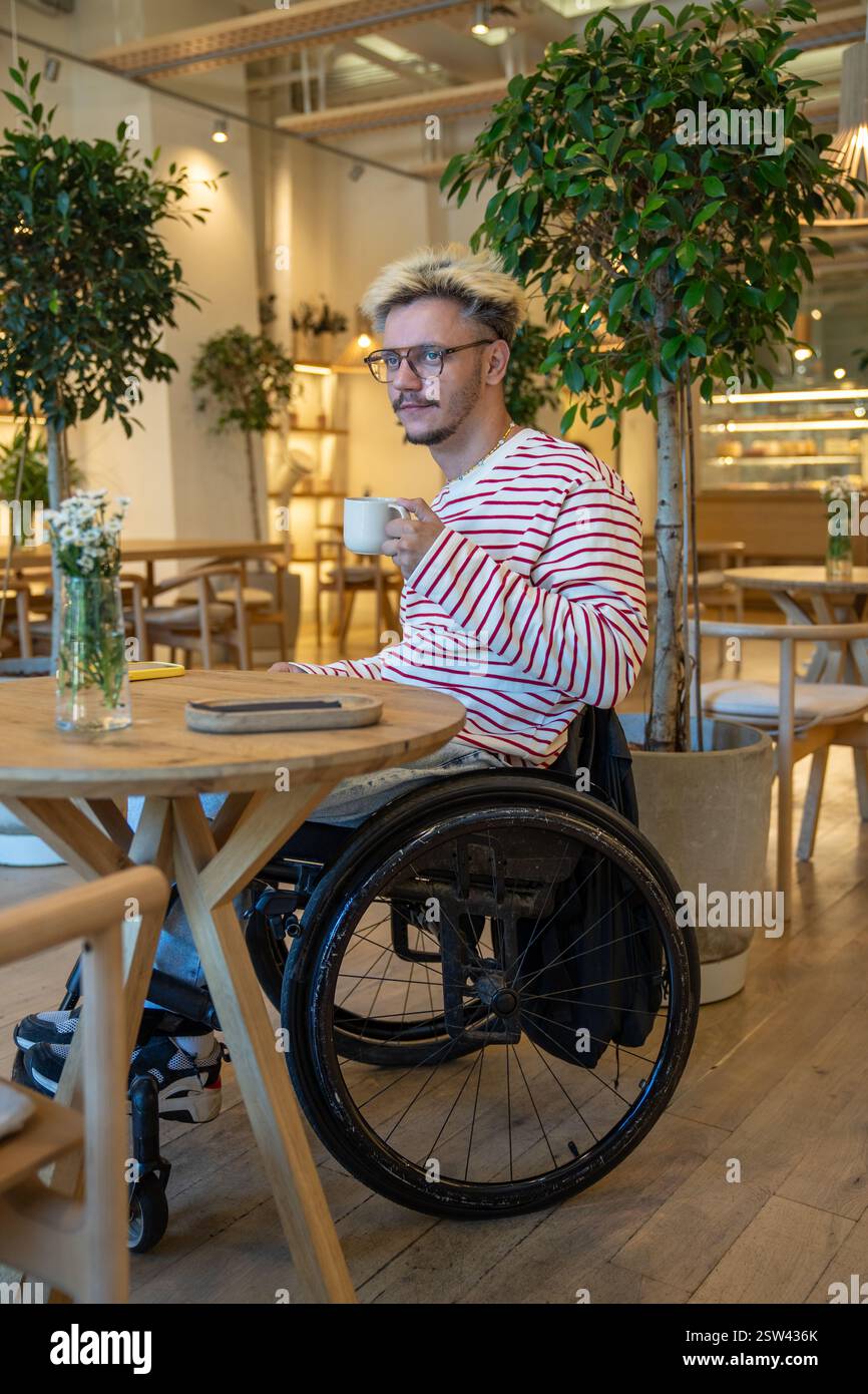 Thoughtful dreamy disabled man in wheelchair drinking coffee at cafe table, waiting for meeting Stock Photo