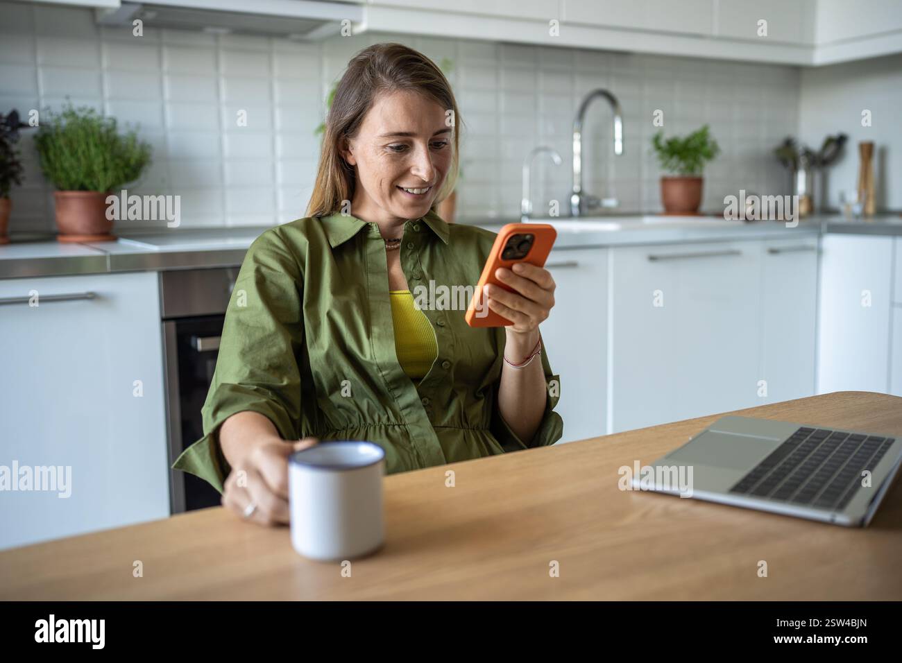 Joyful woman taking break in kitchen, looking on smartphone chatting, scrolling web holding cup tea Stock Photo