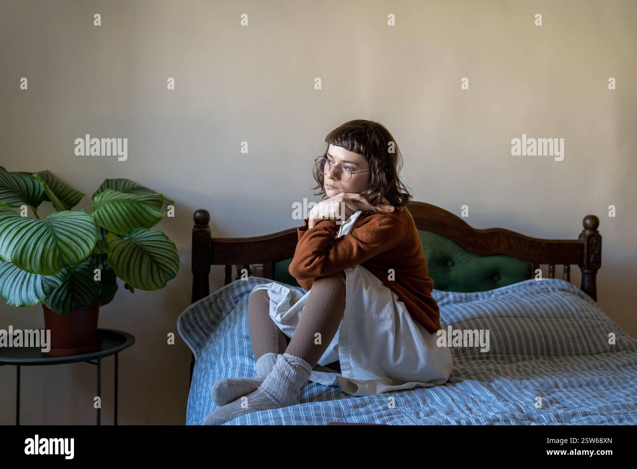 Sad, melancholic woman sits alone on bed, look out window. Feel of loneliness, autumnal melancholy Stock Photo