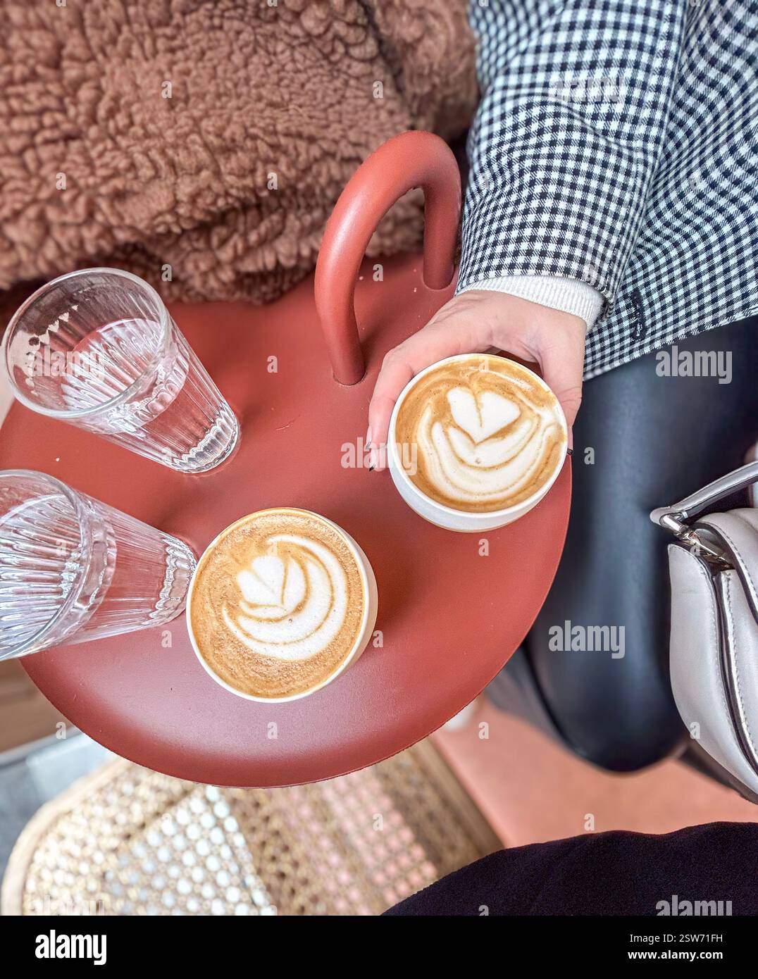 In the centre of Paris, two flat white coffees and two glasses of water on a clay coloured table in a coffee shop. A hand with black tip nails and a b - Smartphone Captured Stock Image