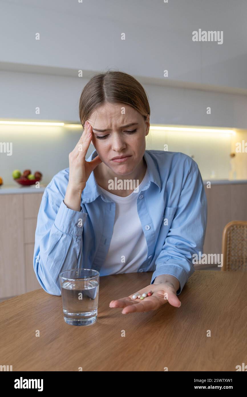 Struggling grimacing woman with headache drinks antidepressants in kitchen. Female mental problems Stock Photo