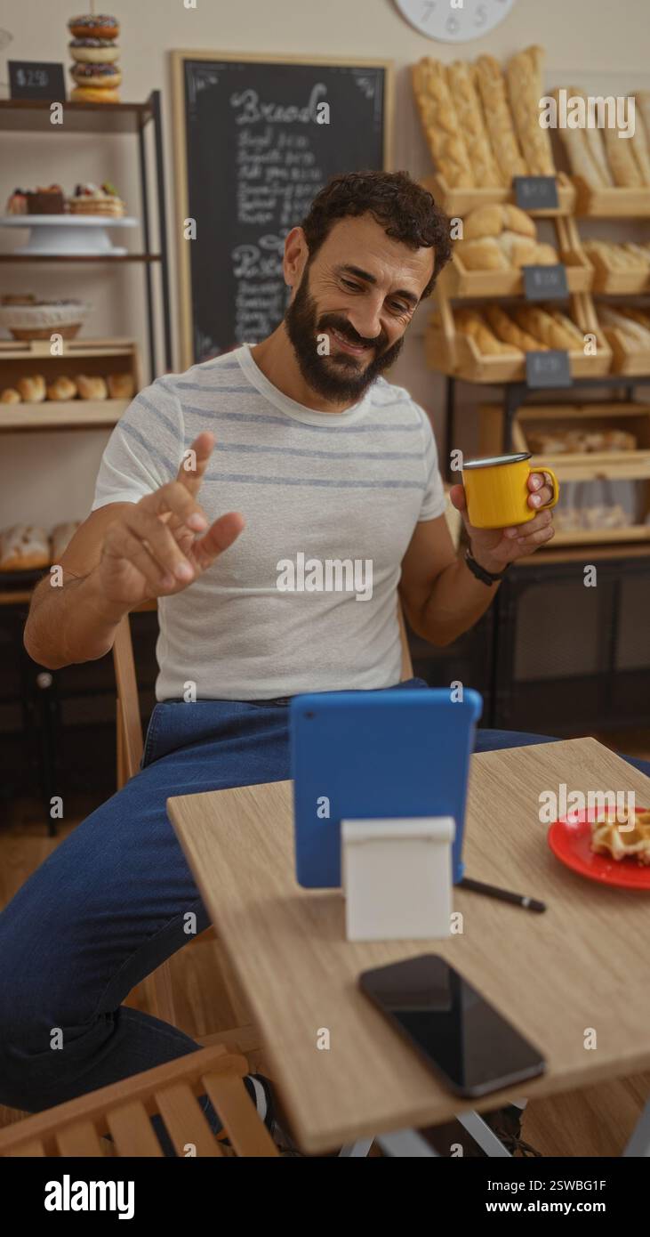 Hispanic man making a video call in a cozy bakery, enjoying a coffee ...