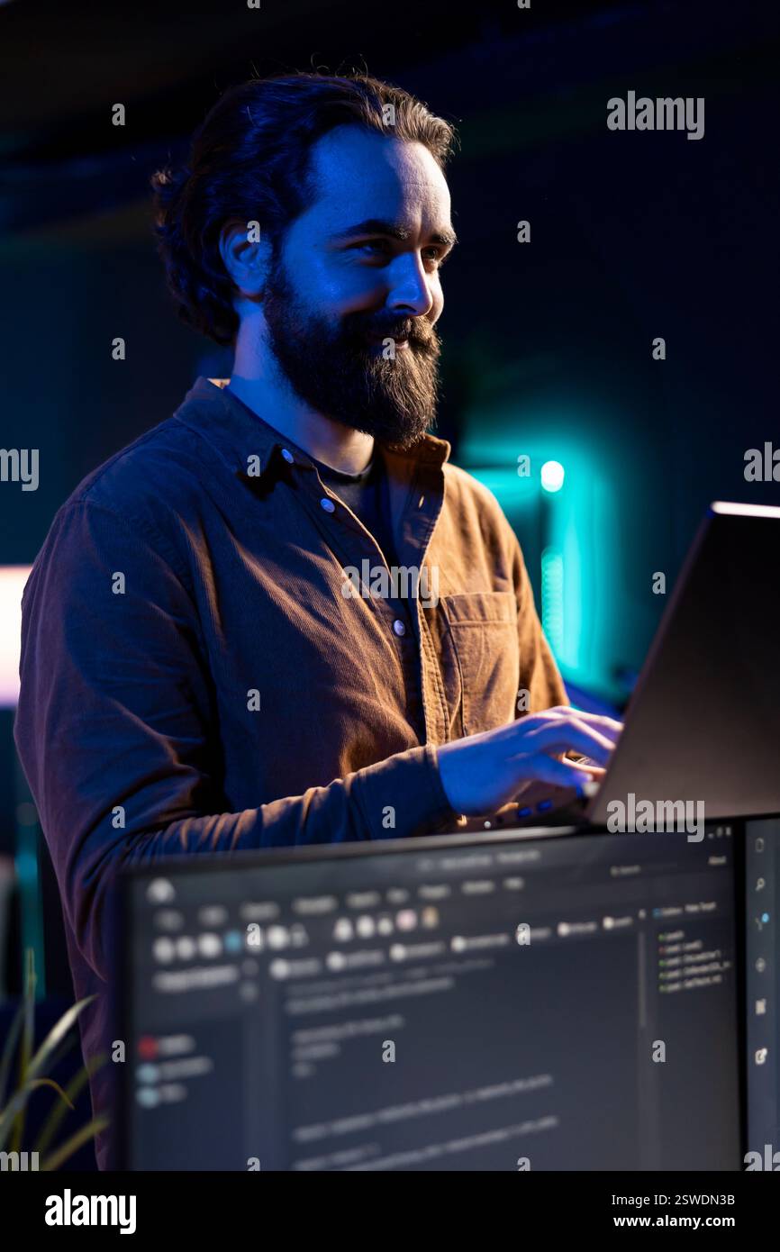 Joyful freelancer doing software debugging using laptop, checking errors in lines of code displayed on PC monitors. Cheerful IT professional walking in apartment fixing coding issues using notebook Stock Photo