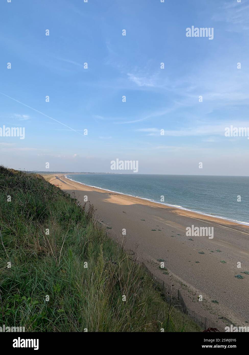 A scenic coastal view with a sandy beach, grassy cliffs, and a calm blue sea under a clear sky. The peaceful shoreline stretches into the distance. - Smartphone Captured Stock Image