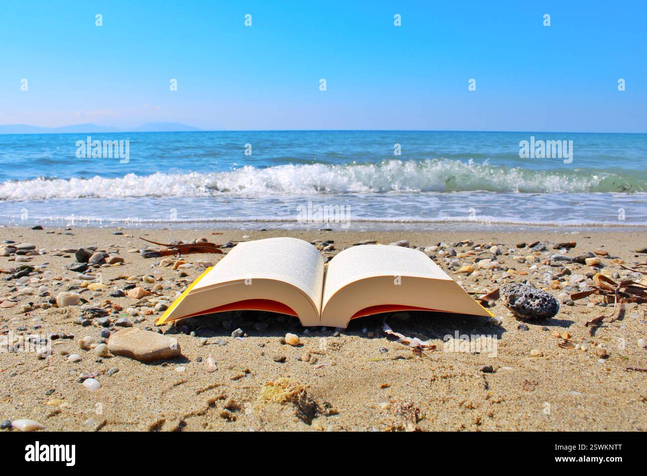 An open book on a sandy beach with pebbles, facing the ocean waves under a clear blue sky. Relaxing summer reading scene by the seaside. Stock Photo
