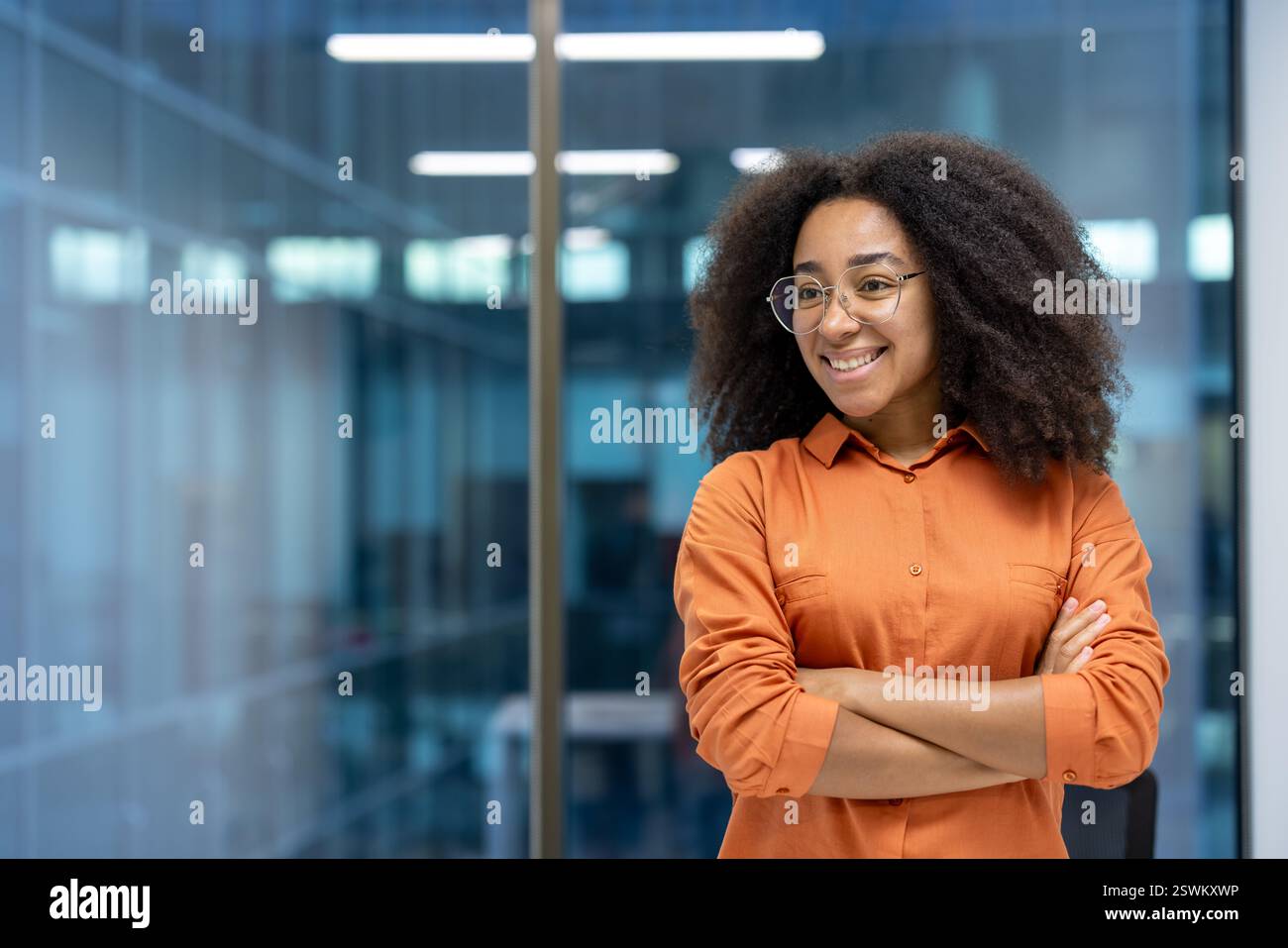Young adult African American woman stands confidently with crossed arms in a modern office setting, smiling and looking away Stock Photo