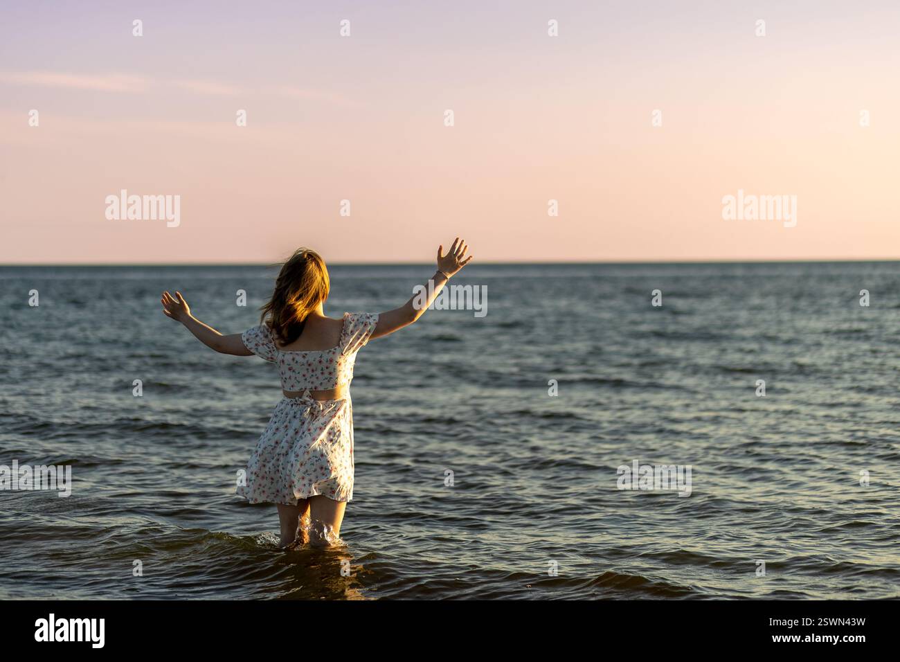 A young woman stands in shallow ocean water with her arms raised ...