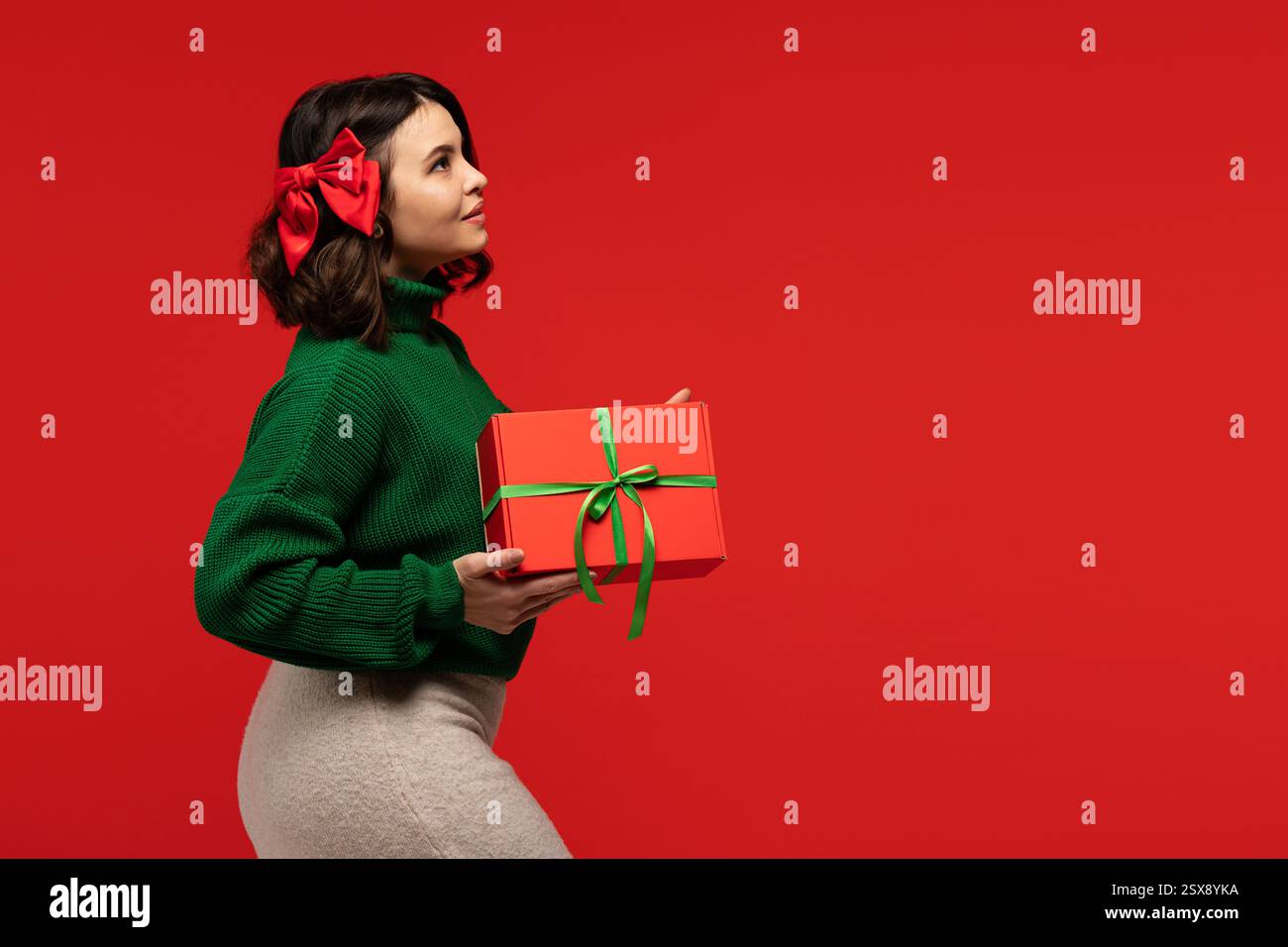 Young woman in a green sweater with a red bow in her hair, holding a wrapped red gift box against a red background. Stock Photo