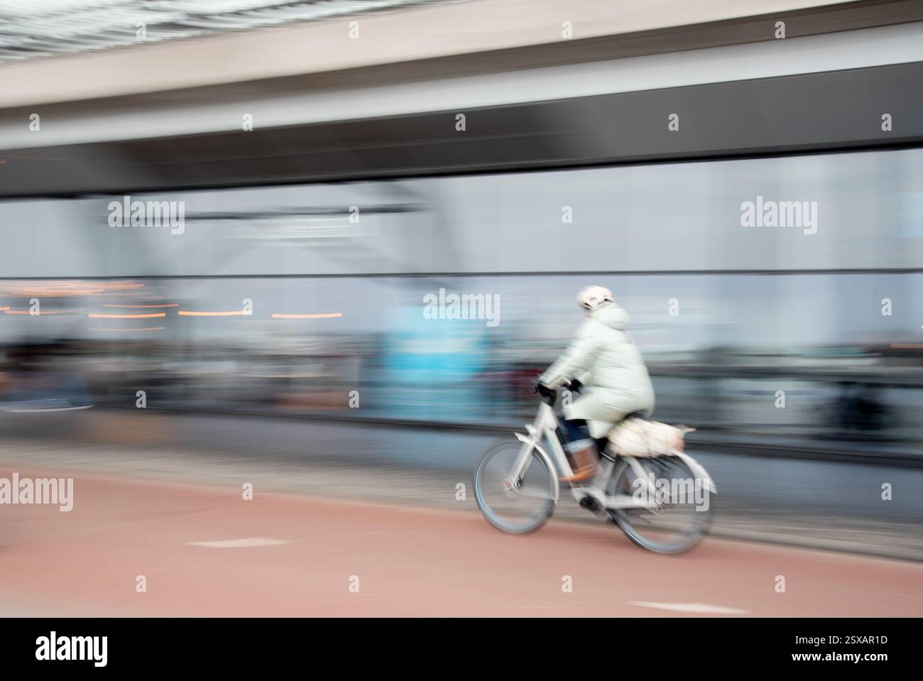 A person dressed in light clothing pedals a bicycle along a paved path in an urban area. The background shows blurred reflections of shops and people, Stock Photo
