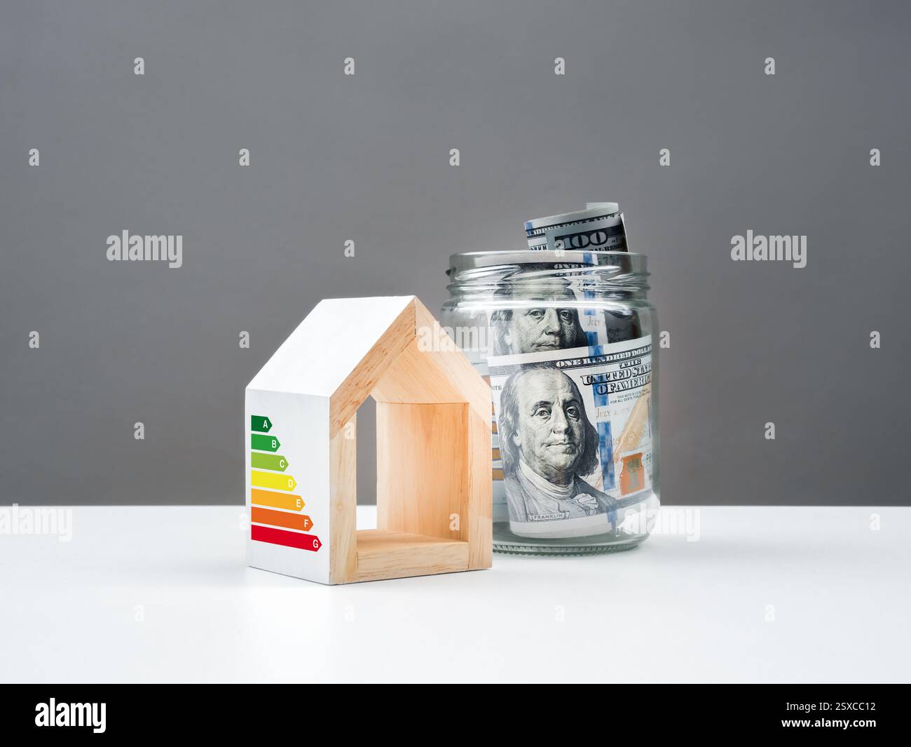 Efficient energy concept, sustainable energy efficiency saving home. One hundred dollars bill banknotes in glass jar near white house model with rate Stock Photo