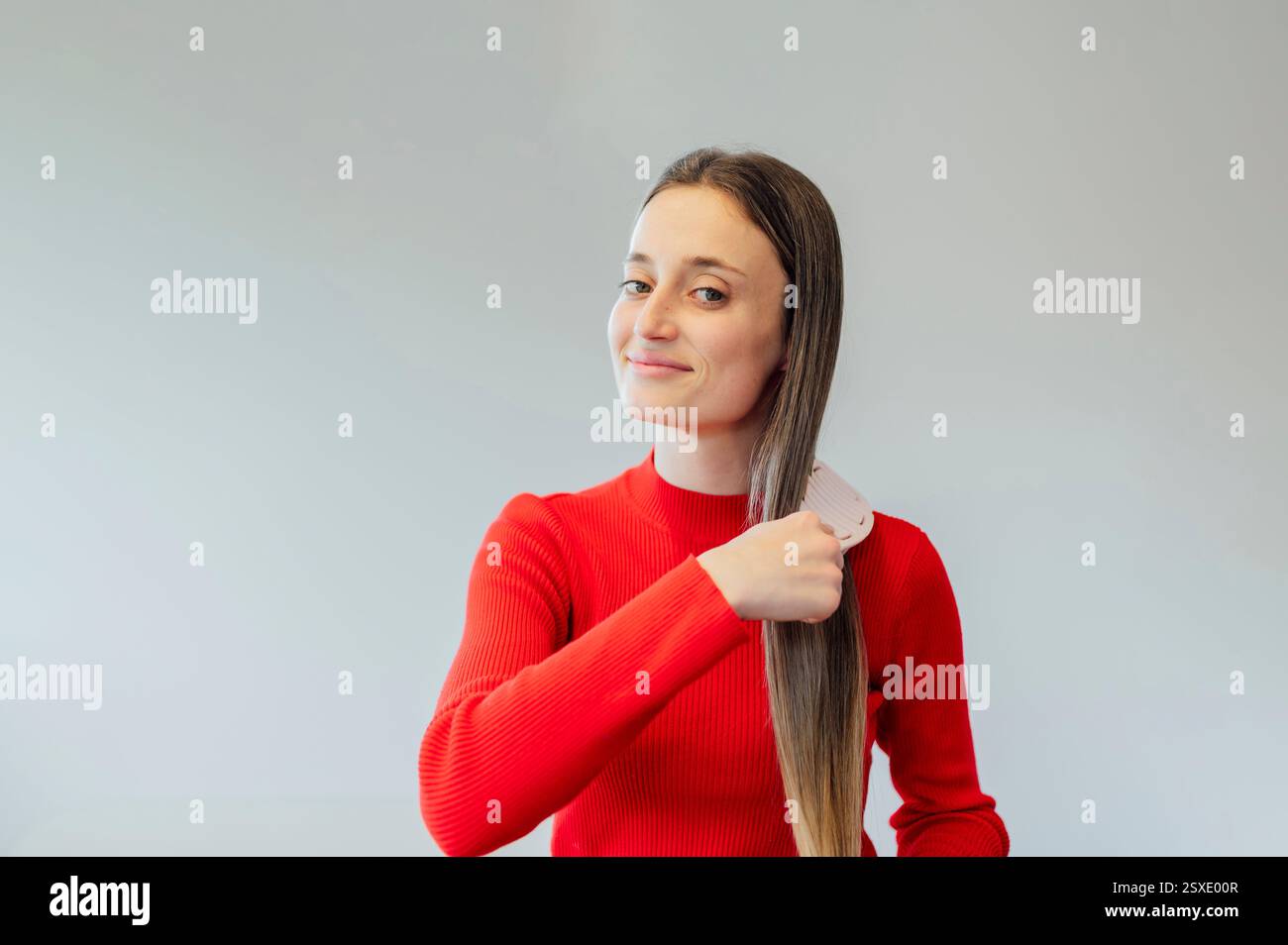 Young woman combing her long hair with comb, wearing red sweater Stock Photo