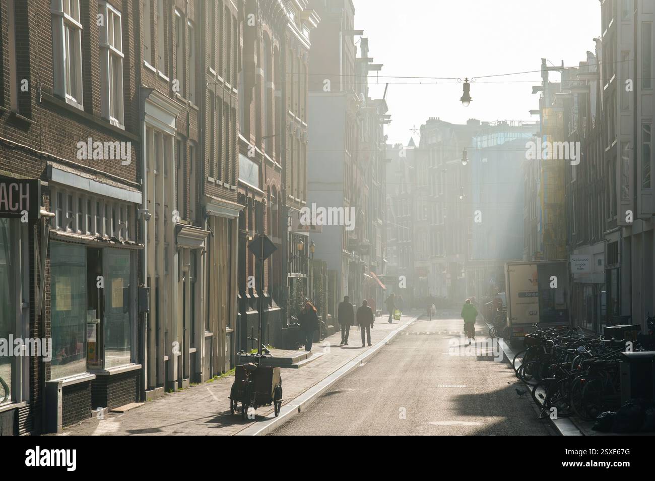 A sunlit morning street in Amsterdam with historic buildings, bicycles, and pedestrians walking through soft mist, creating a serene urban atmosphere. Stock Photo