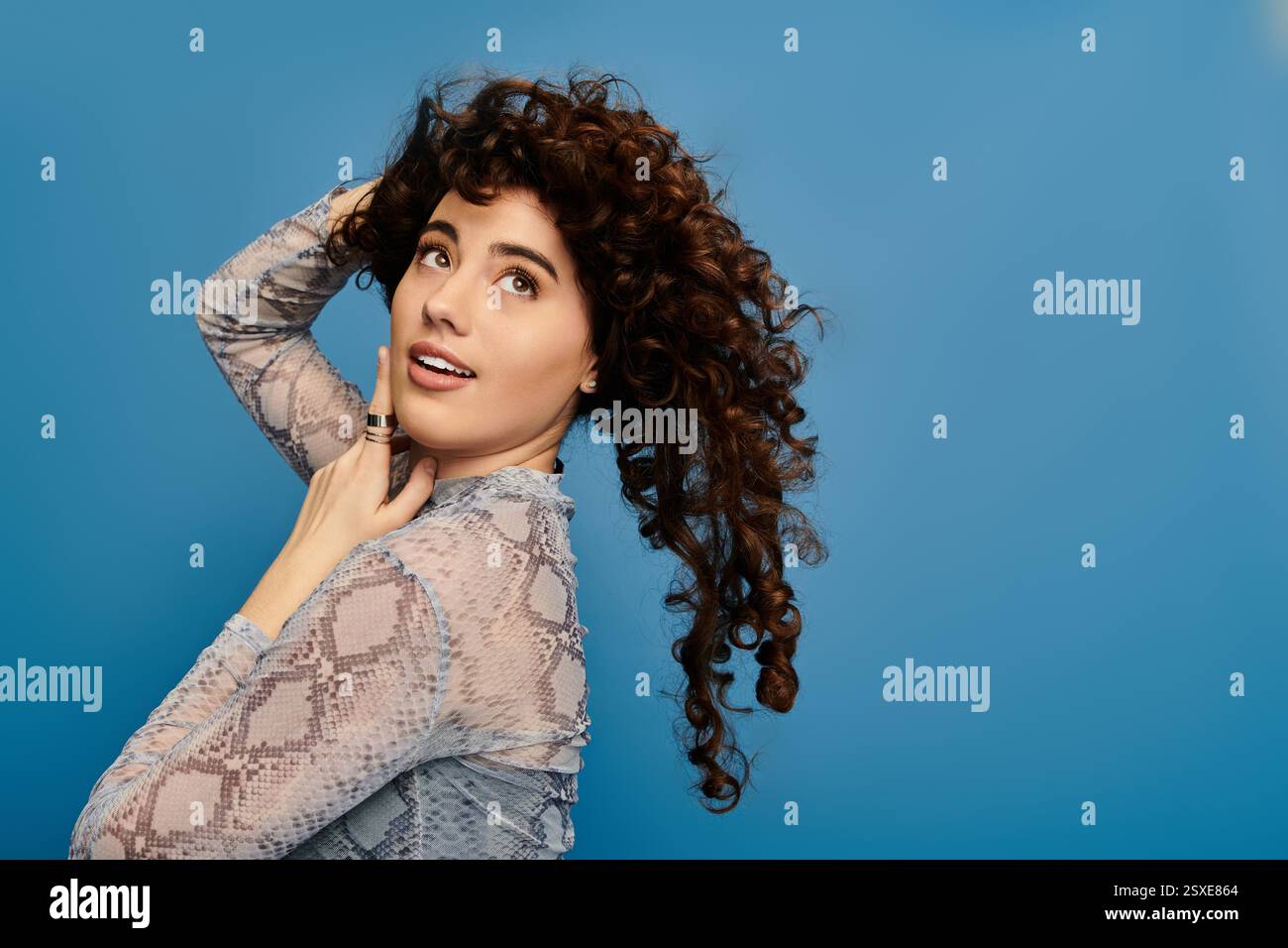 Emotional woman with curly hair poses playfully by a blue wall ...