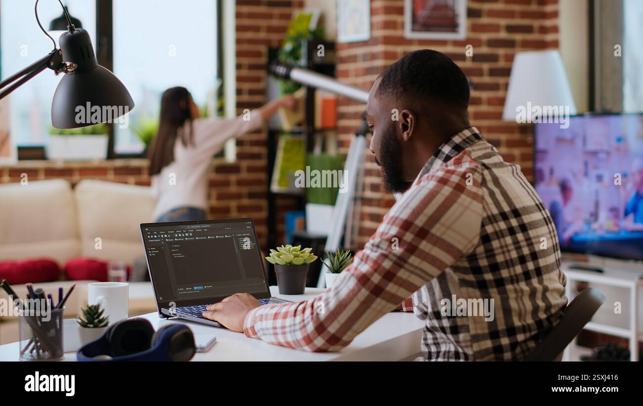 African american software developer works on programming code on a laptop in remote workspace. Productive IT engineer working on coding, software design and internet security innovation. Camera B. Stock Photo
