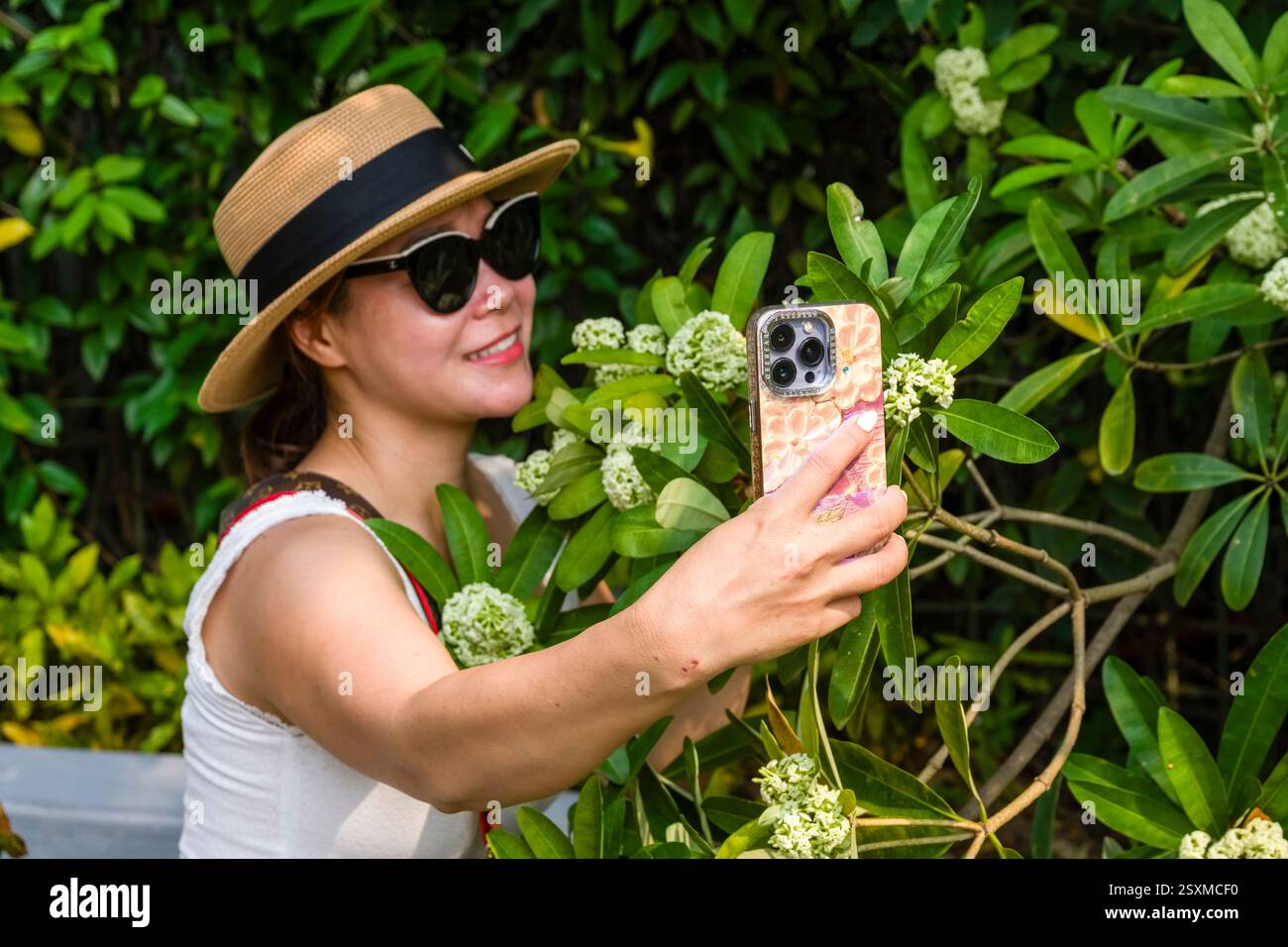 A smiling young woman wearing a sun hat and sunglasses, surrounded by bushes of blossoming flowers, takes a selfie with her mobile phone. Stock Photo
