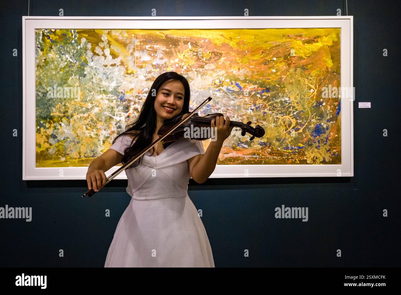 A smiling young woman in a white dress plays the violin in front of an abstract-style painting. Stock Photo