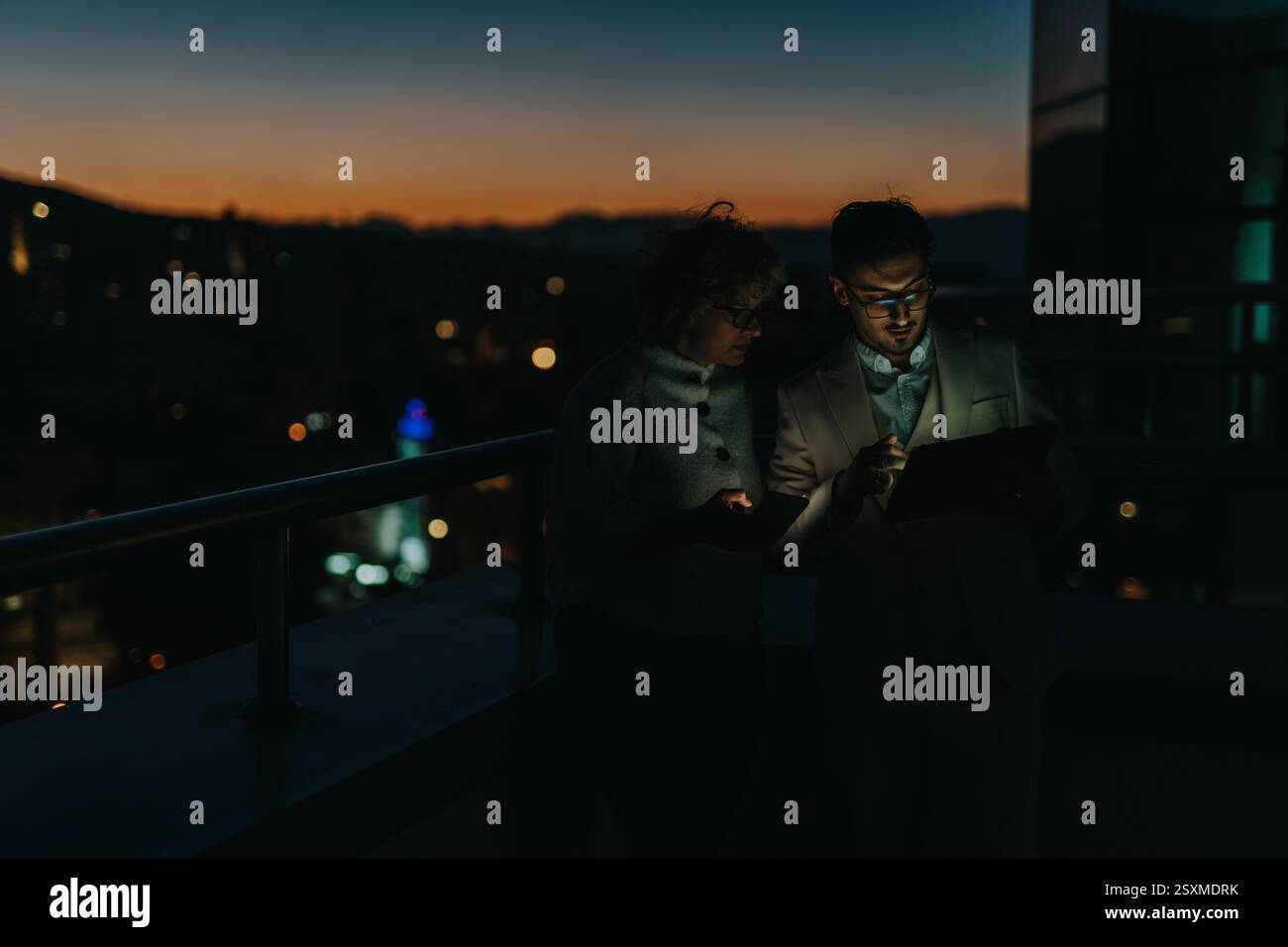 Two business people collaborate outdoors at night, using a tablet for brainstorming ideas. Stock Photo