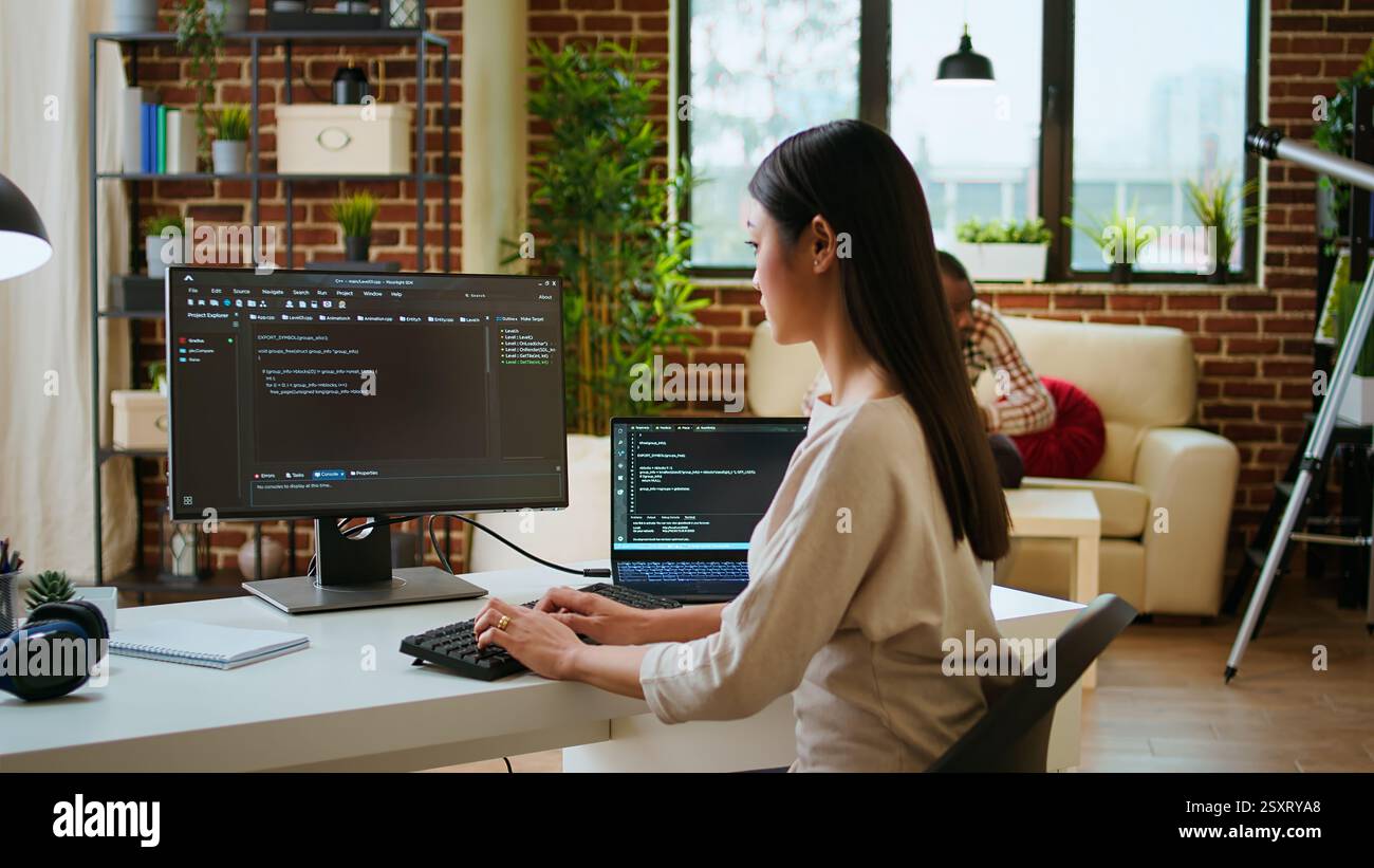 Asian programmer seen typing code for a user interface design on dual monitors, working in a digital workspace. Woman coder working on software engineering and front end development. Camera A. Stock Photo