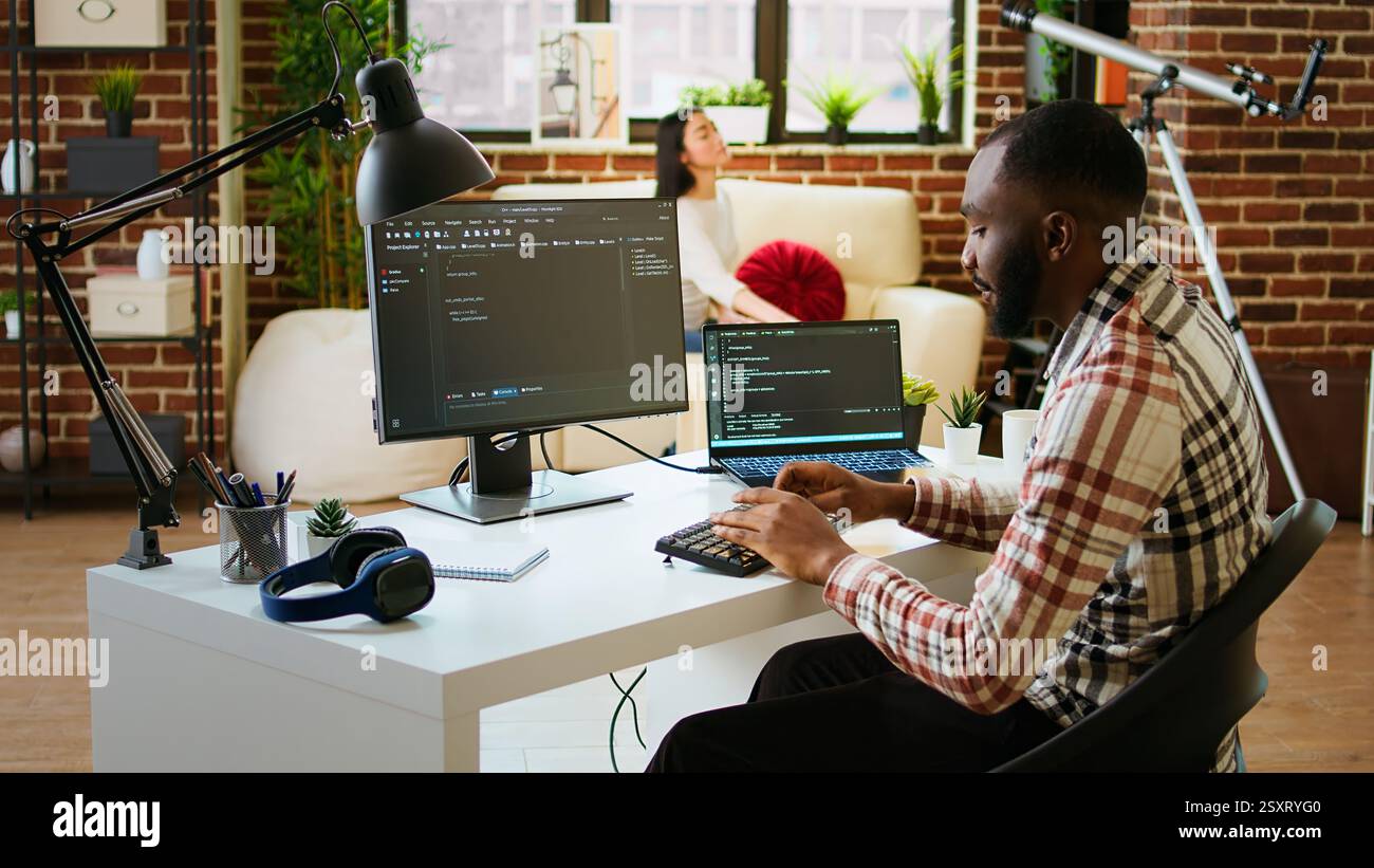 Young man seen programming software code on his laptop in a sleek home office. Workspace with dual monitors helps with software engineering, web development and coding productivity. Camera A. Stock Photo