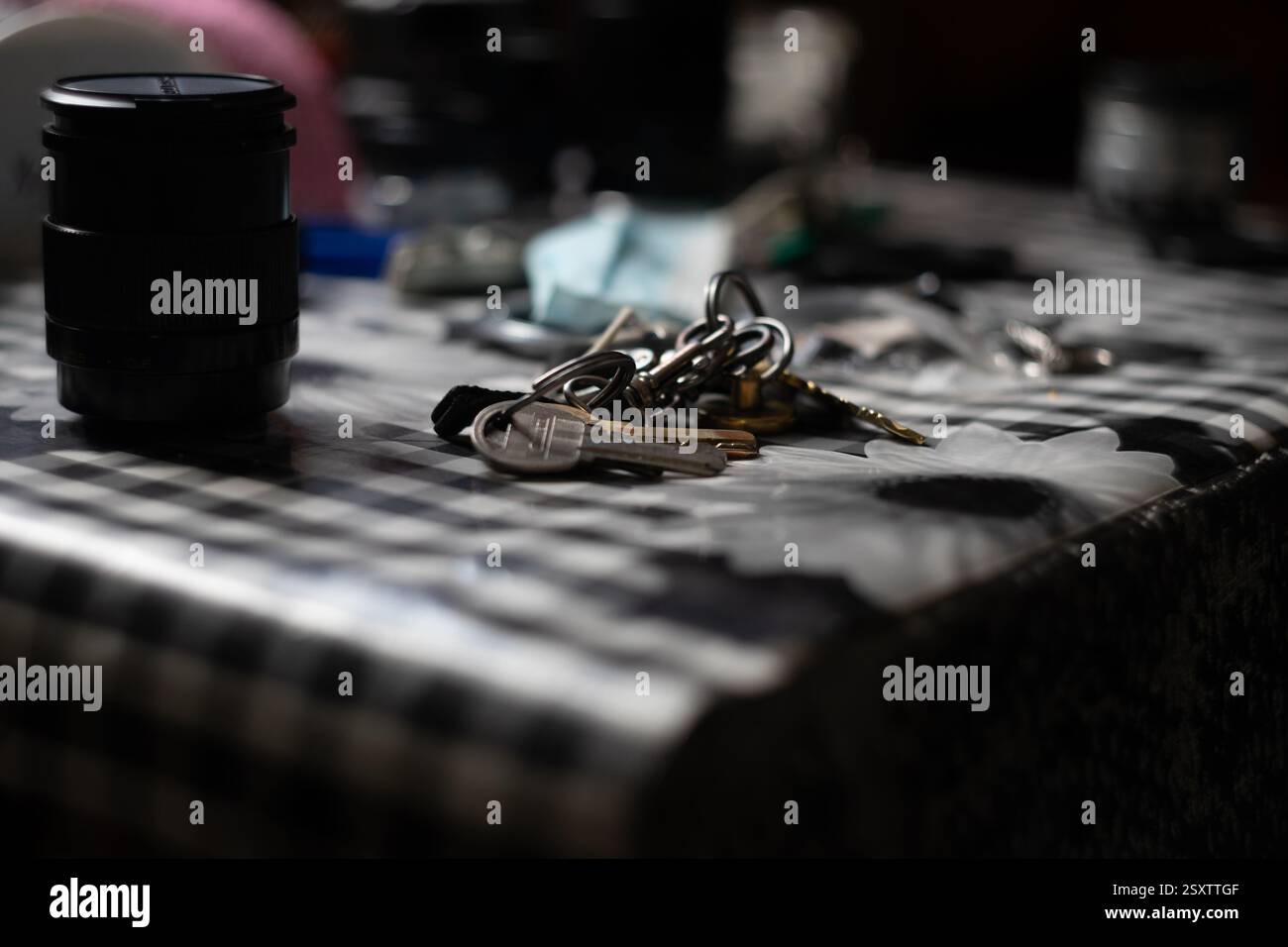 Keys rest among scattered items on a checkered tablecloth. A camera lens sits nearby, surrounded by various objects in a softly lit room, creating a c Stock Photo