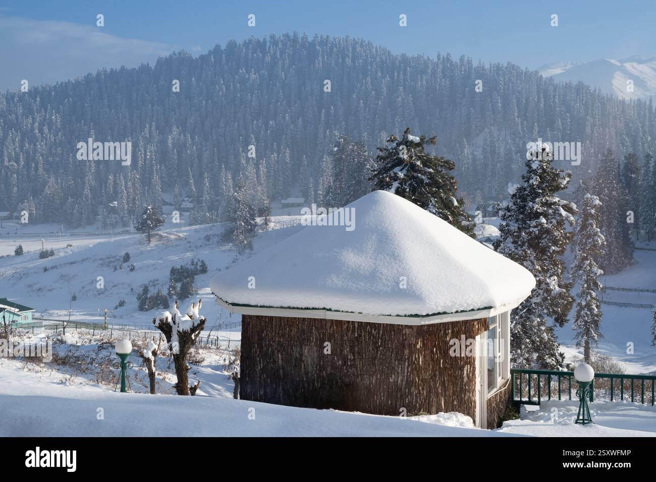 wooden cabins covered in snow with pine deodar trees surrounding it and the Himalaya mountains peeking out of the fog in the distance in Gulmarg Stock Photo