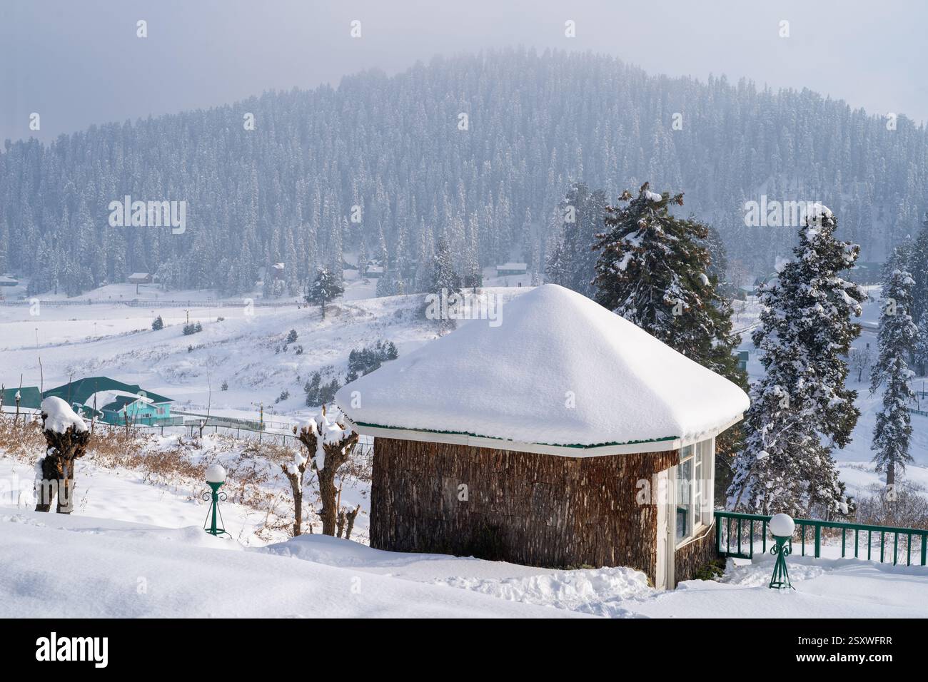 wooden cabins covered in snow with pine deodar trees surrounding it and the Himalaya mountains peeking out of the fog in the distance in Gulmarg Stock Photo