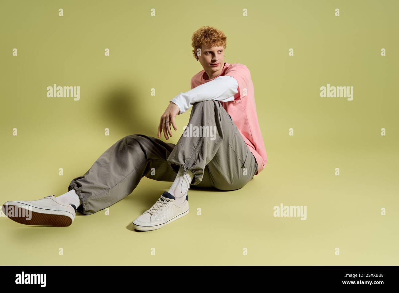 A fashionable young man with curly hair relaxes in a stylish outfit on a smooth pastel surface. Stock Photo