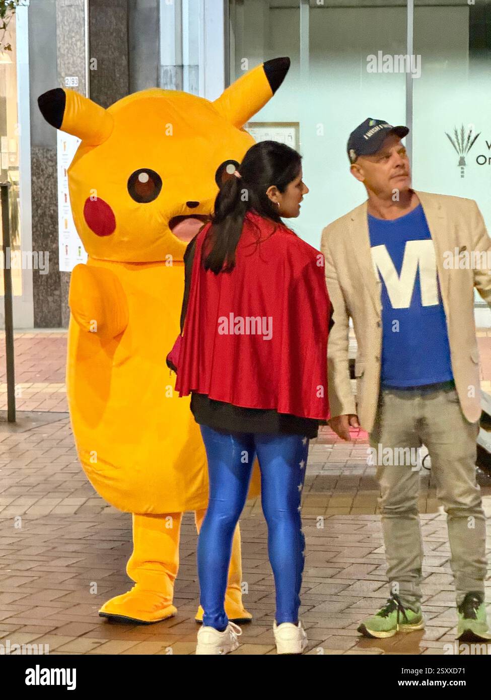 Couple standing next to a person wearing Pokemon Pikachu character costume in the street - Smartphone Captured Stock Image