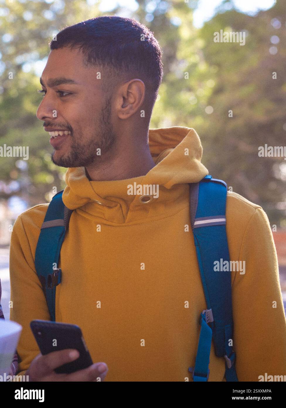 Smiling young man in yellow hoodie holding smartphone outdoors with backpack Stock Photo