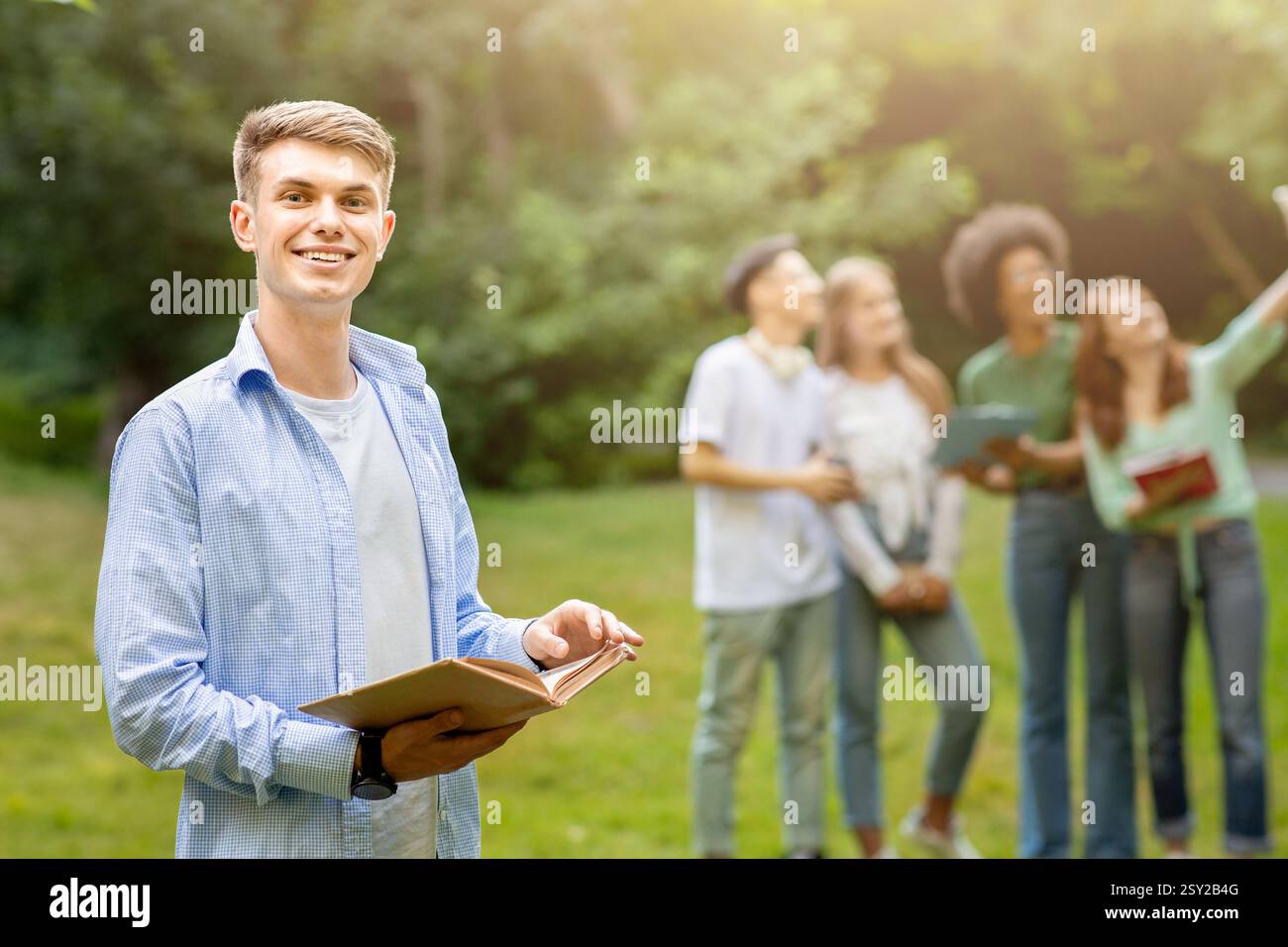 Education First. Student Guy Posing With Book While His Classmates Taking Selfie Stock Photo