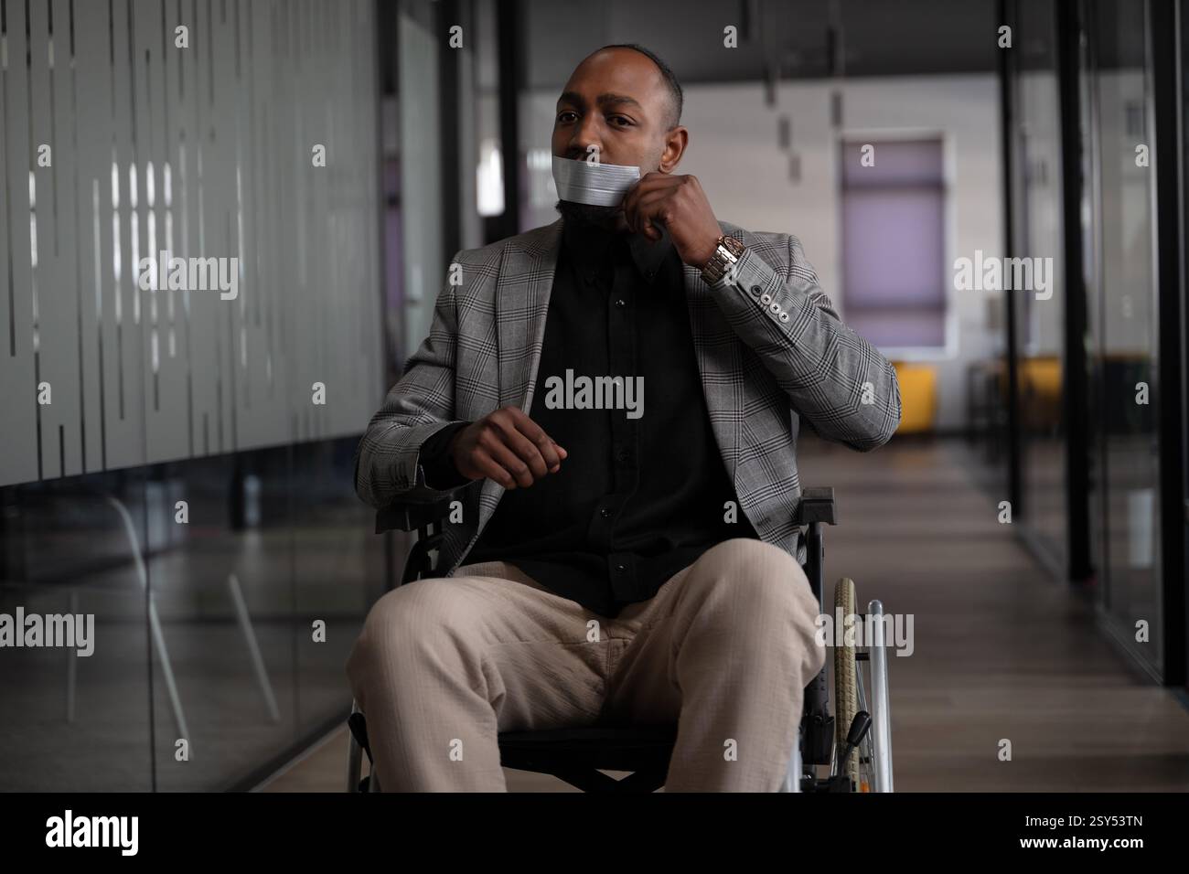 African man in a wheelchair wearing a stylish blazer and removing duct tape from his mouth in a modern office corridor. Concept of freedom of speech Stock Photo