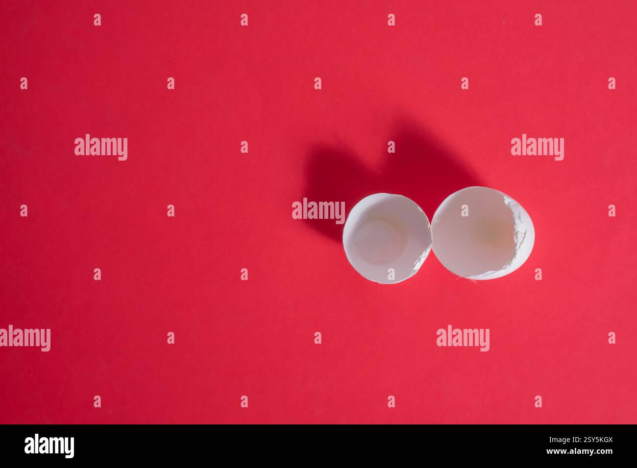 Two empty eggshell halves are set against a vibrant red surface, casting soft shadows Stock Photo