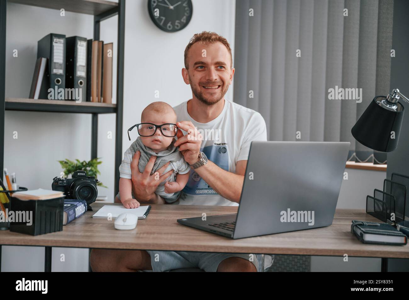 Big glasses. Sitting by the table with laptop. Father with his newborn son is indoors at home. Stock Photo