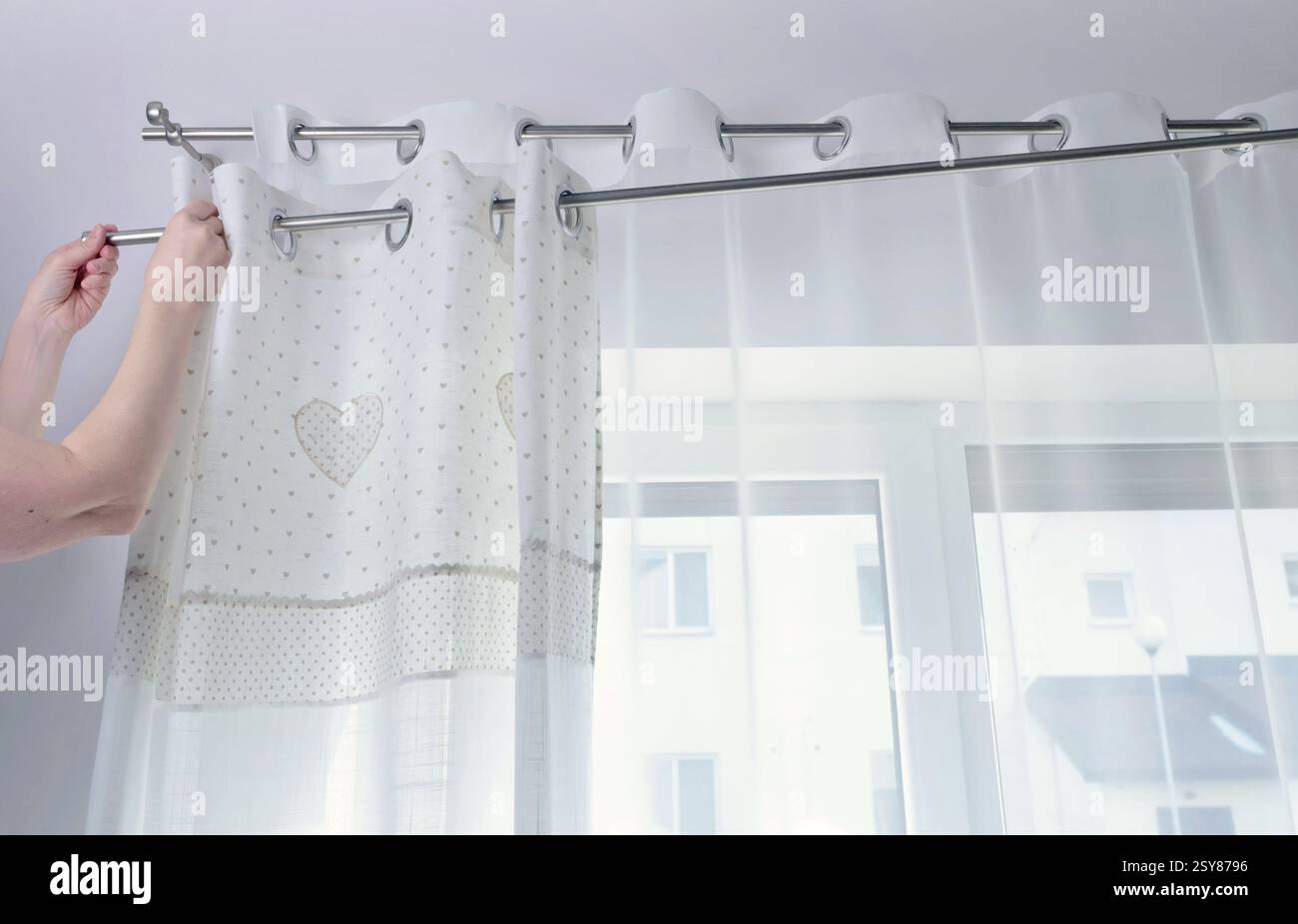 Woman hangs white curtains with metal rings made of natural material with embroidered hearts in kitchen of house. Cleaning and cleanliness. Interior Stock Photo